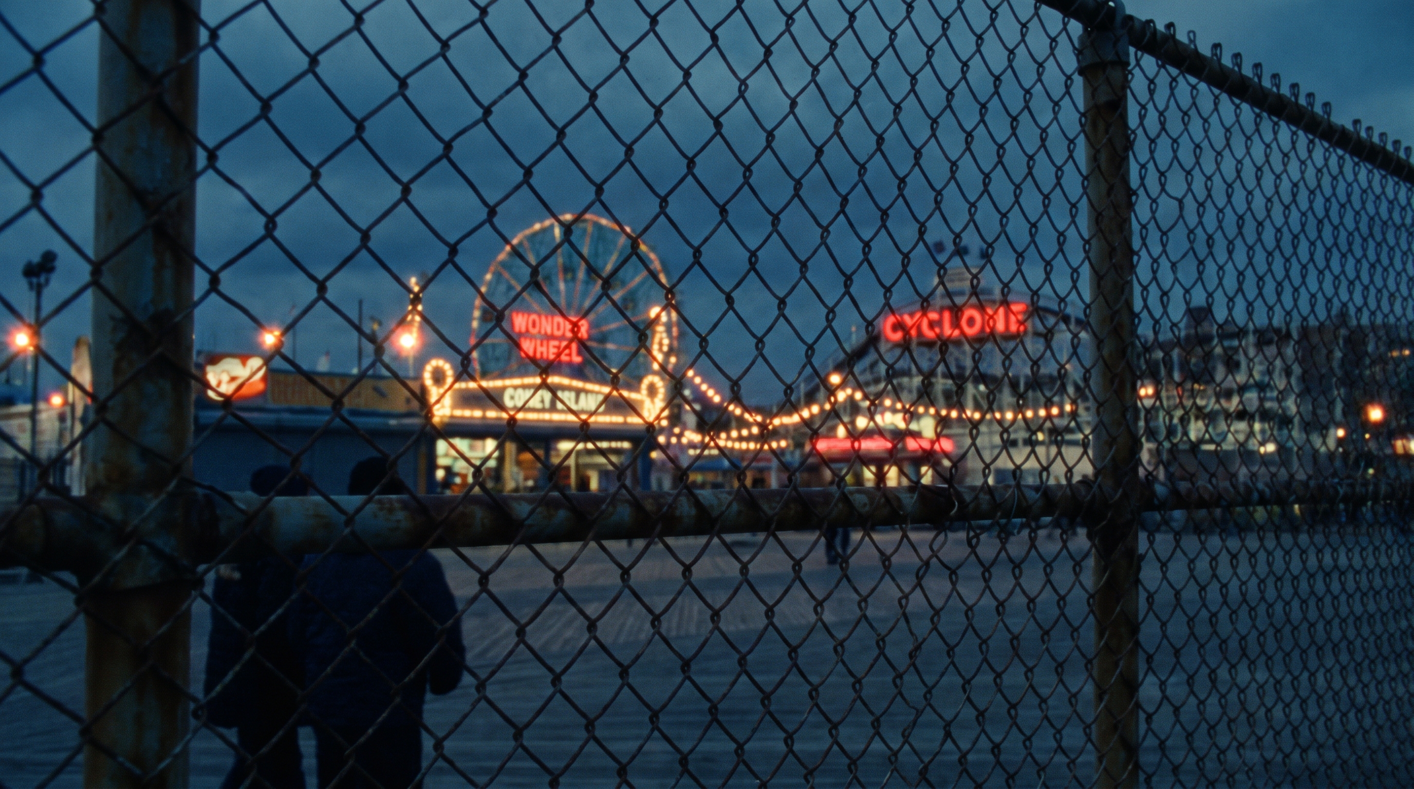The Coney Island amusement district seen at dusk through a chain-link fence, the Wonder Wheel and Cyclone lit in red neon, two bundled figures walking the near-empty boardwalk.