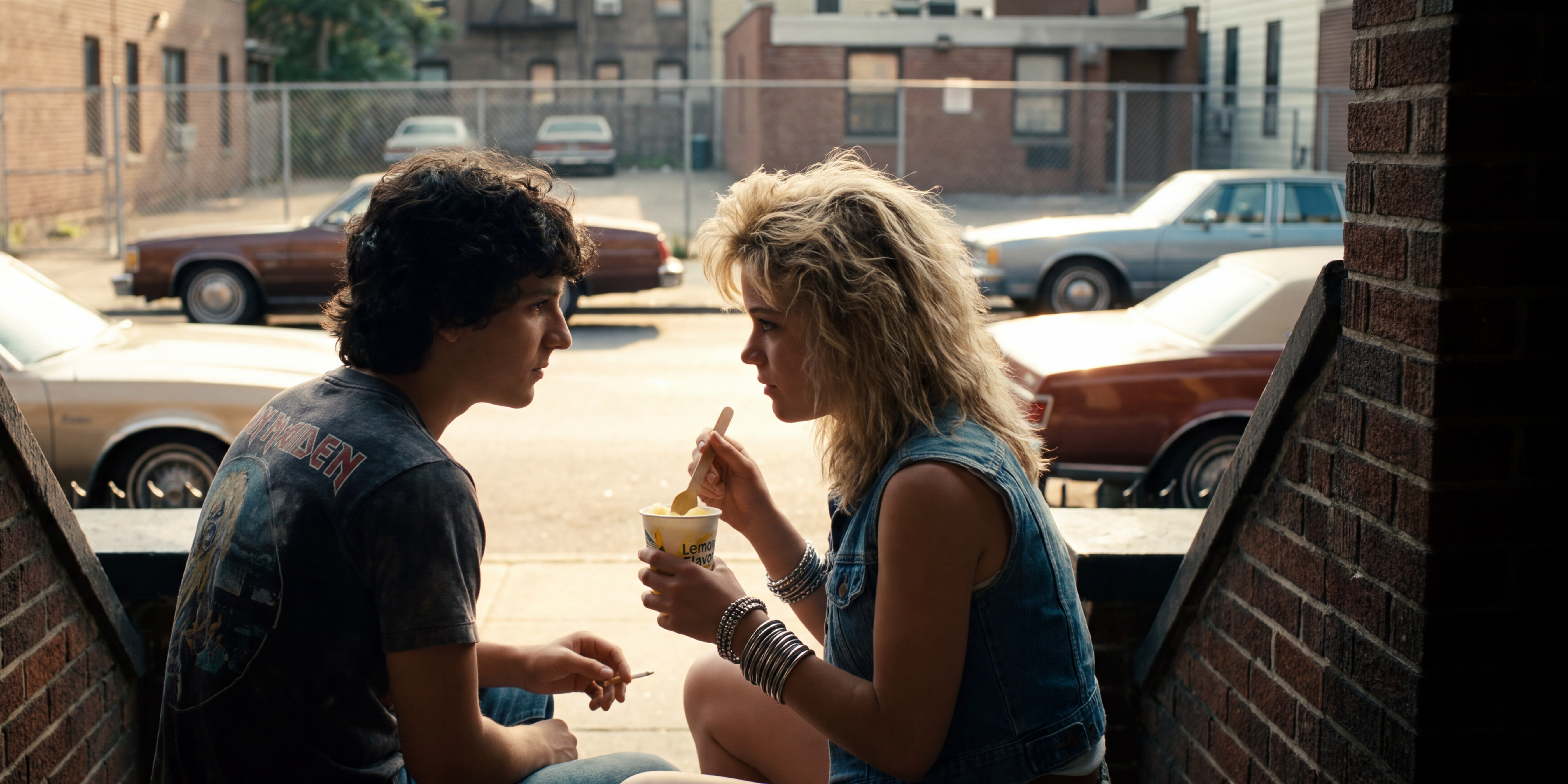 A teenage boy in a Maiden t-shirt and a blonde girl in double denim sit close on a Brooklyn brownstone stoop in golden light, sharing a paper cup of lemon ice with a small wooden spoon, 1980s sedans parked along the street.